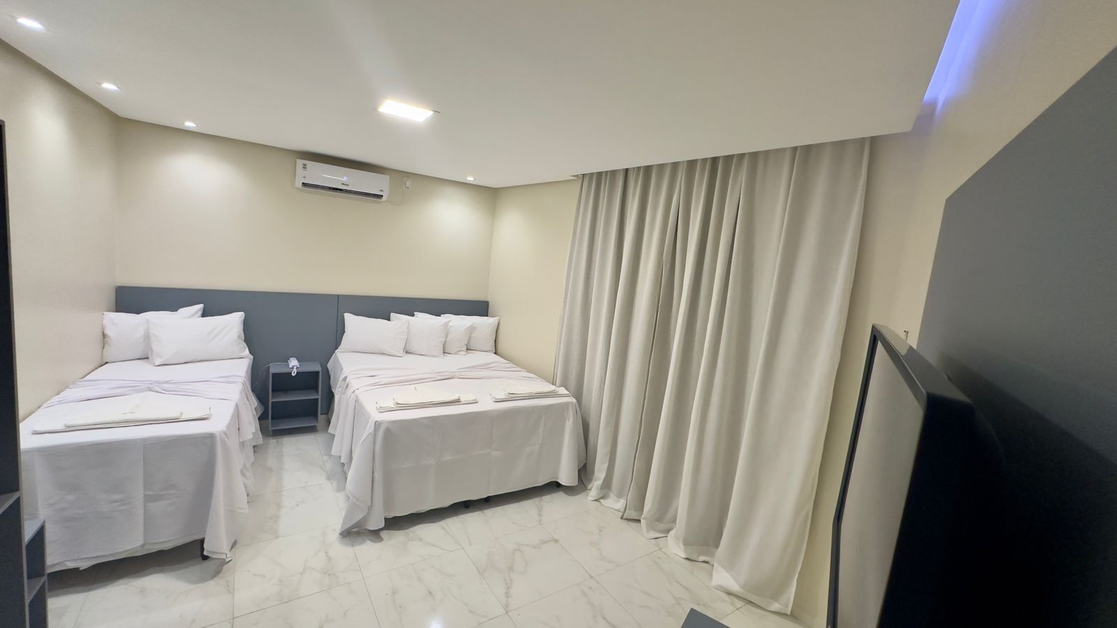 A hotel room with two beds, a flat-screen TV, a bedside table, and a large window with curtains.