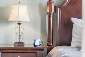 Ein ordentlich bezogenes Bett mit weißen Leinen, ein Nachttisch aus Holz mit Lampe und Tasse sowie ein Holz-Kopfteil.