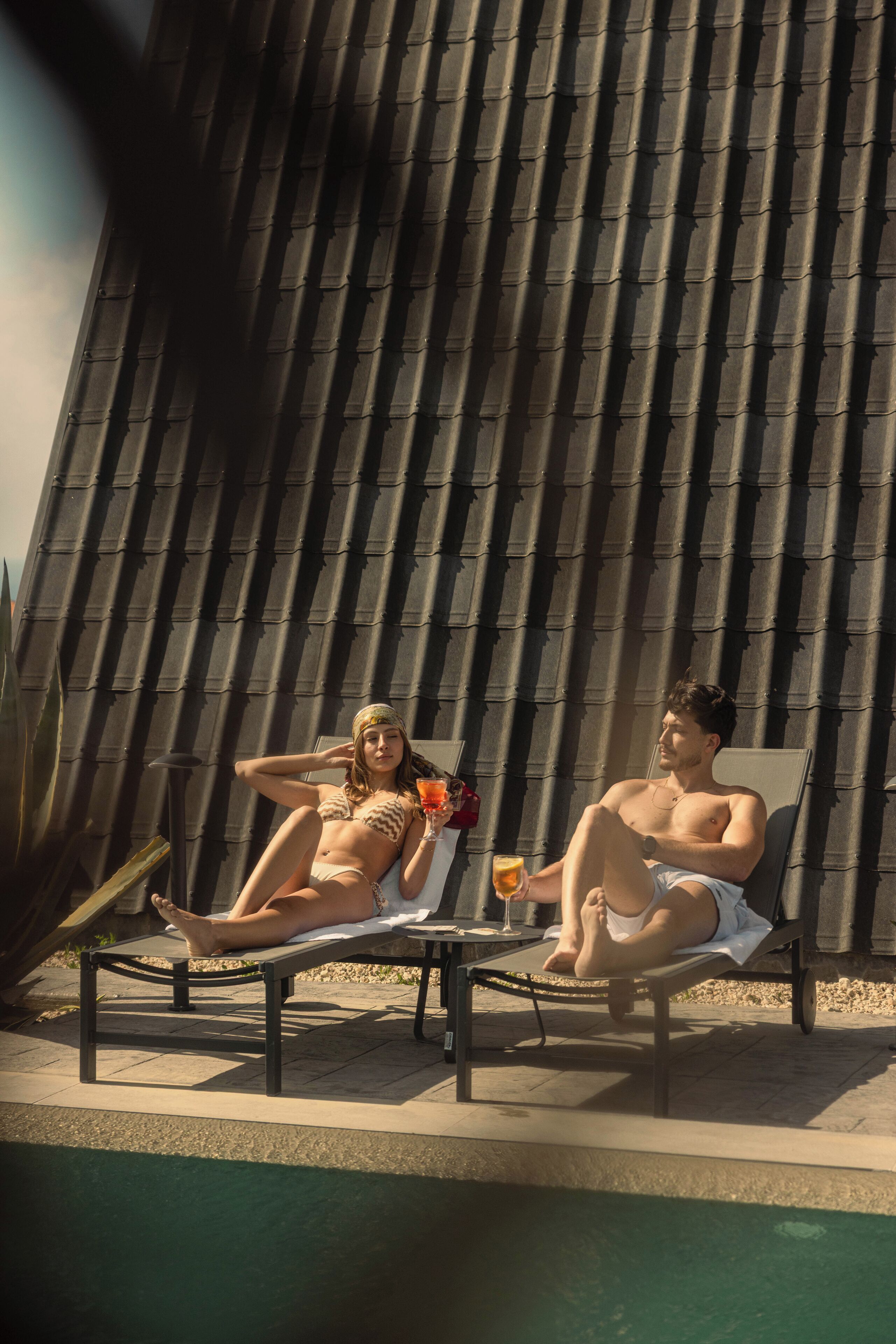 Two people relaxing on lounge chairs by a pool with a tiled wall in the background.