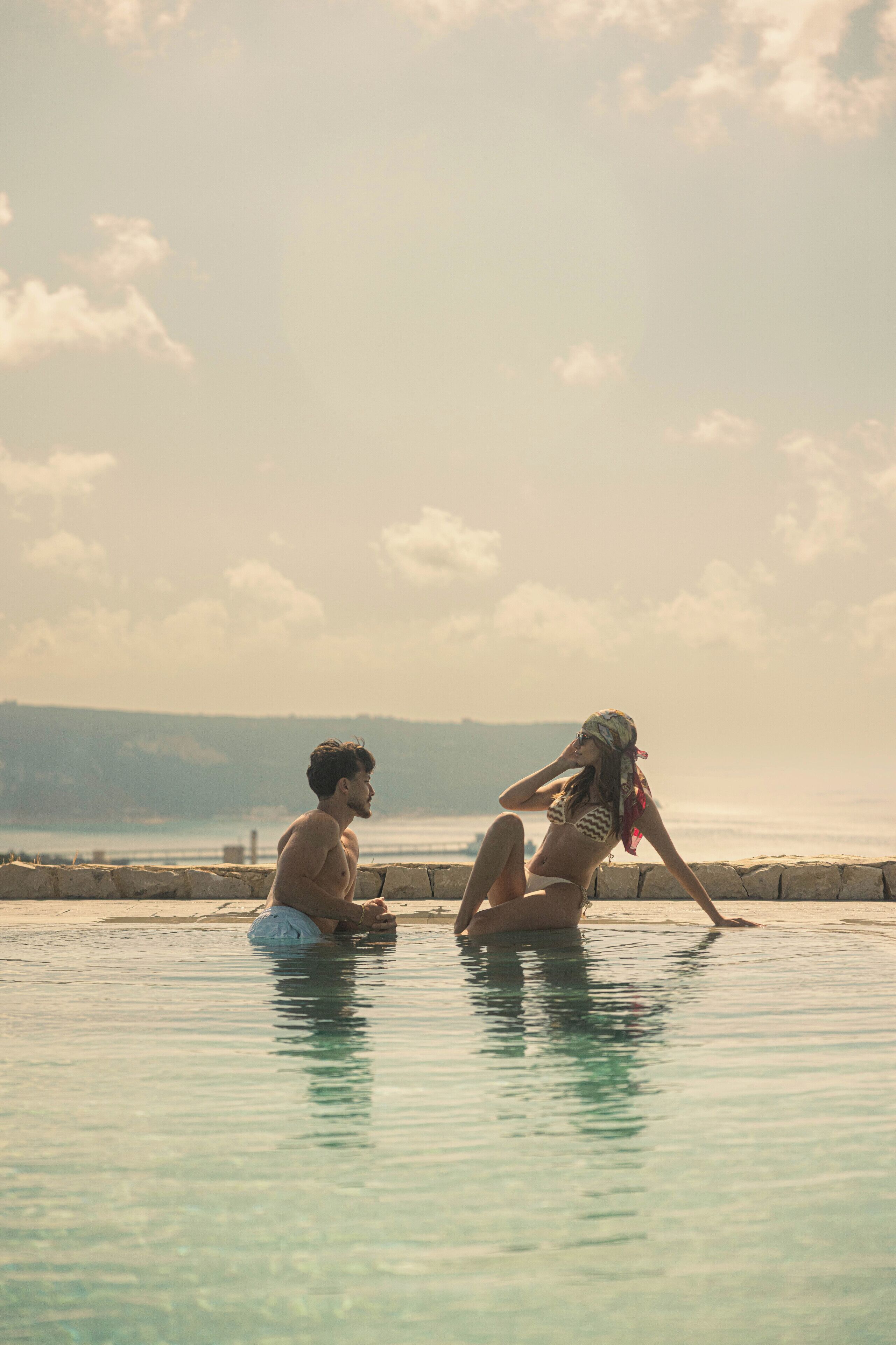 A man and a woman sitting by a pool with a view of the sea and hills in the background.