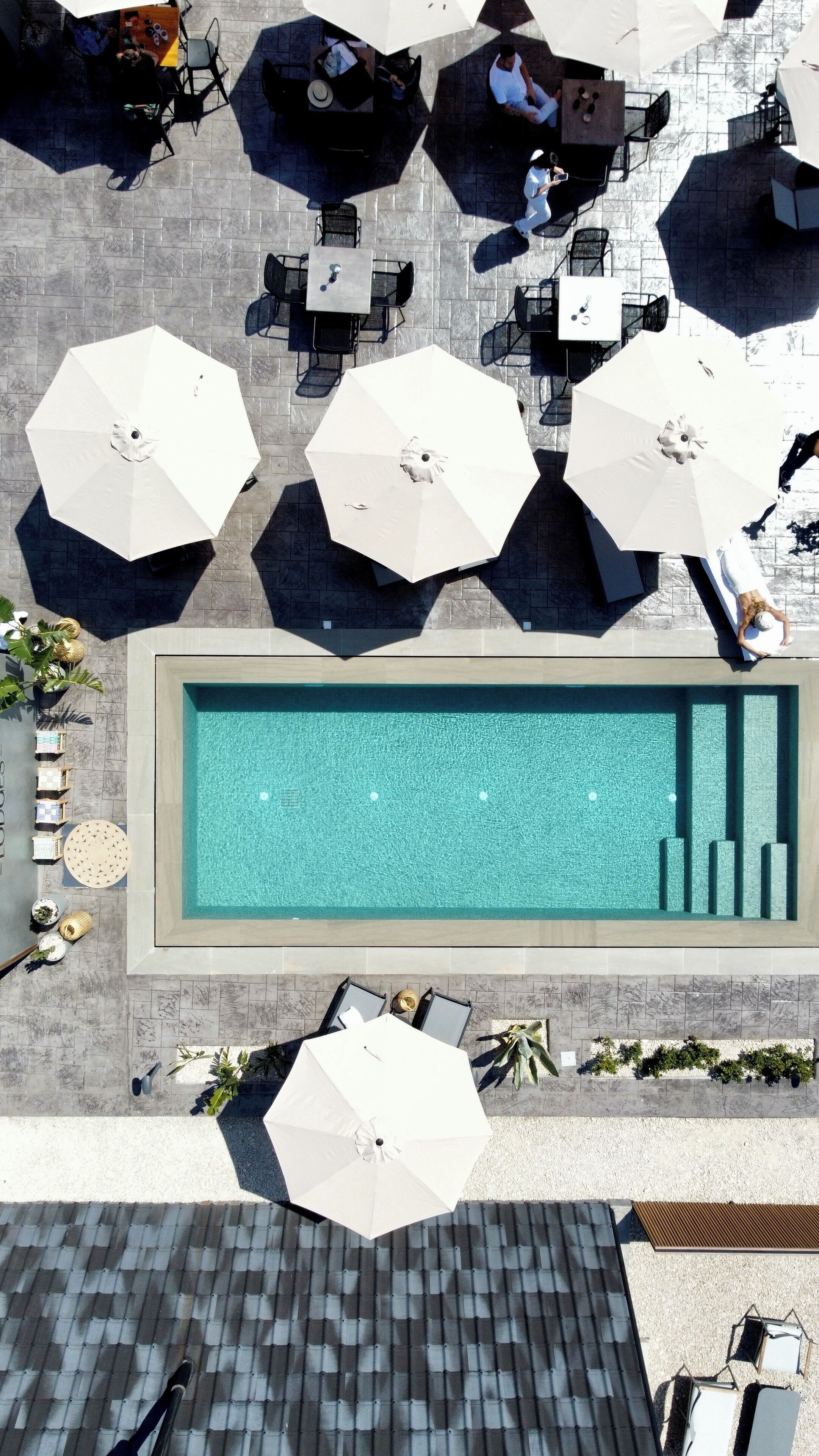 Vue aérienne d’un espace piscine sur le toit, avec plusieurs parasols blancs, des chaises longues et une piscine.