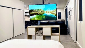 A modern hotel room with a large flat-screen TV mounted on the wall, a bed with a white duvet, and a wooden bedside table with a folded blanket. - Modern Studio Home SolMod # 1 (Tucson)