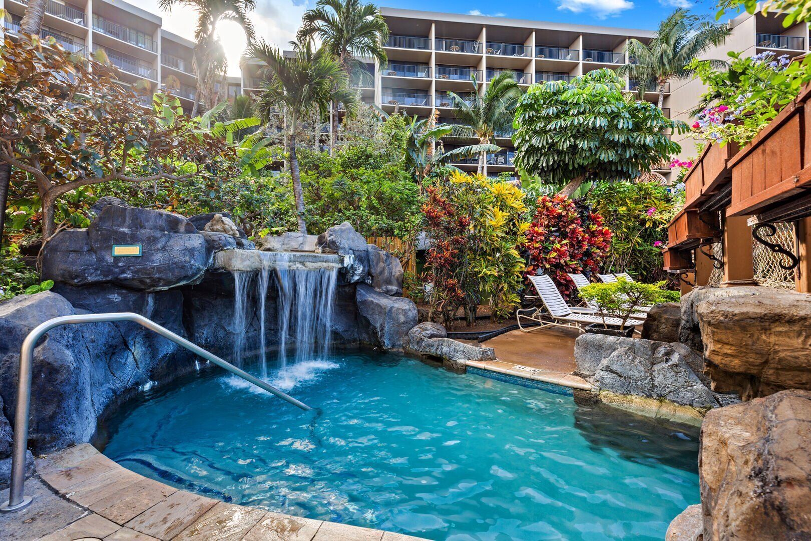 A pool with a waterfall, surrounded by tropical plants and palm trees, with a view of hotel buildings in the background.