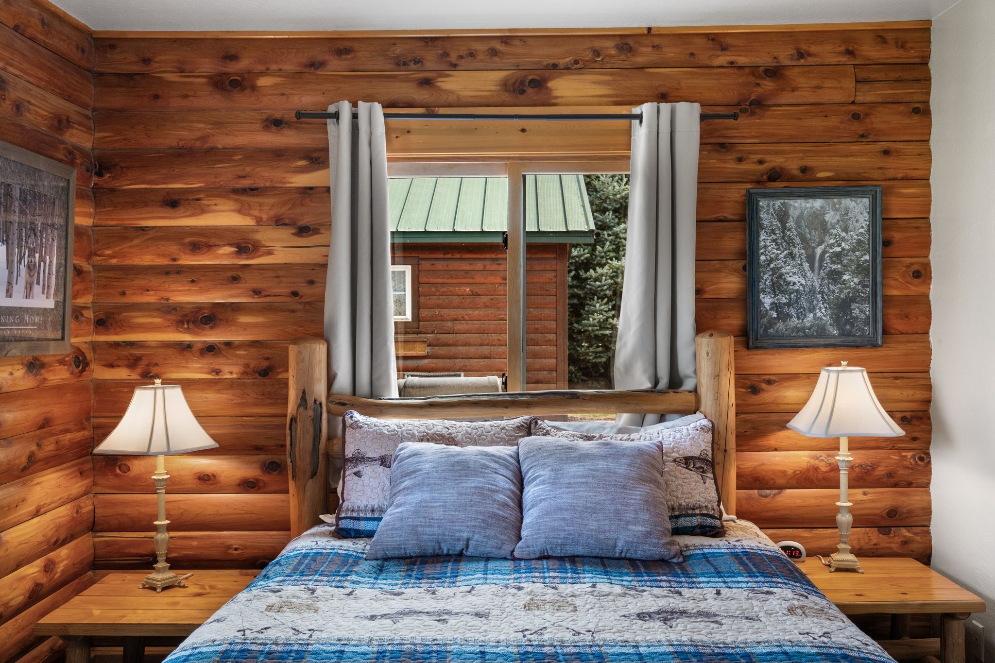 A wooden cabin-style bedroom with a bed, two bedside lamps, a window with curtains, and a framed picture on the wall.