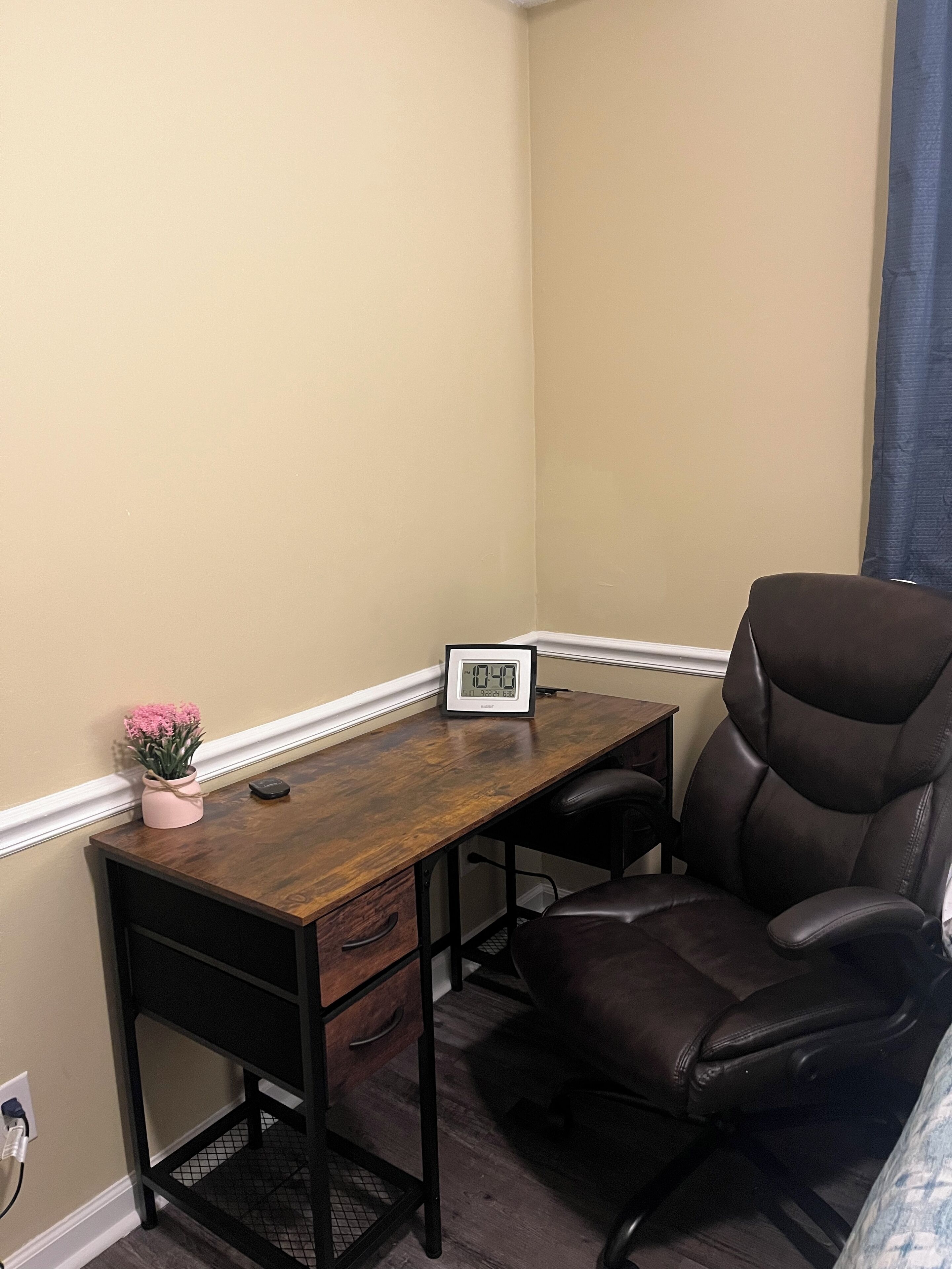 Un bureau d’angle avec un bureau en bois, un fauteuil en cuir noir, une petite plante en pot et une horloge numérique.