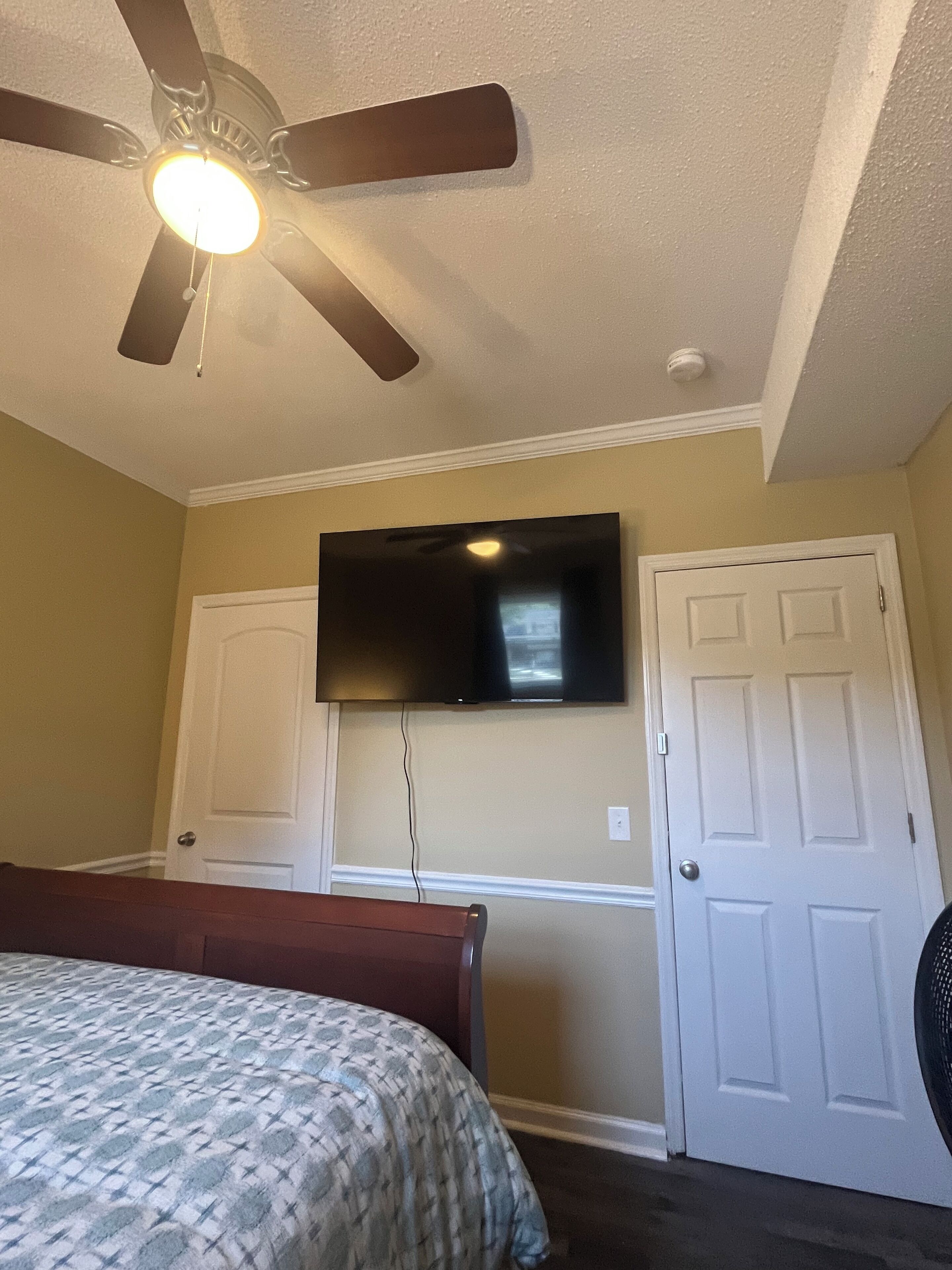 Un dormitorio con cama, ventilador de techo, televisor de pantalla plana montado en la pared y dos puertas blancas.