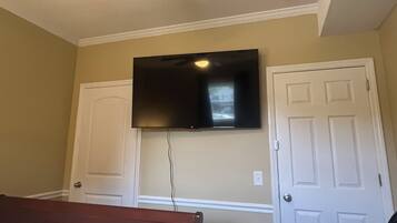 Un dormitorio con cama, ventilador de techo, televisor de pantalla plana montado en la pared y dos puertas blancas.