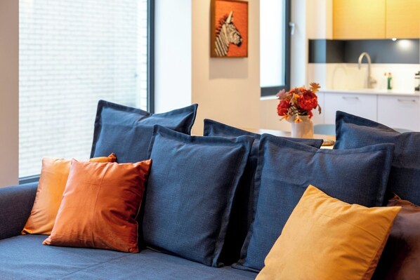 A modern living room with a sofa and multiple cushions, a kitchen area with a sink and cabinets, and a window with blinds.
