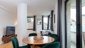 A modern living room with a round wooden table, green velvet chairs, a flat-screen TV, and large windows offering a view of the city. - Modern Flat with Private Balcony | Lift access (Greater London)
