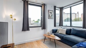 A modern living room with a dark blue sofa, a wooden coffee table, and a large window with curtains. - Modern Flat with Private Balcony | Lift access (Greater London)