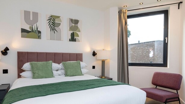A neatly made bed with a green throw, a red chair, and abstract art on the wall. - Modern Flat with Private Balcony | Lift access (Greater London)