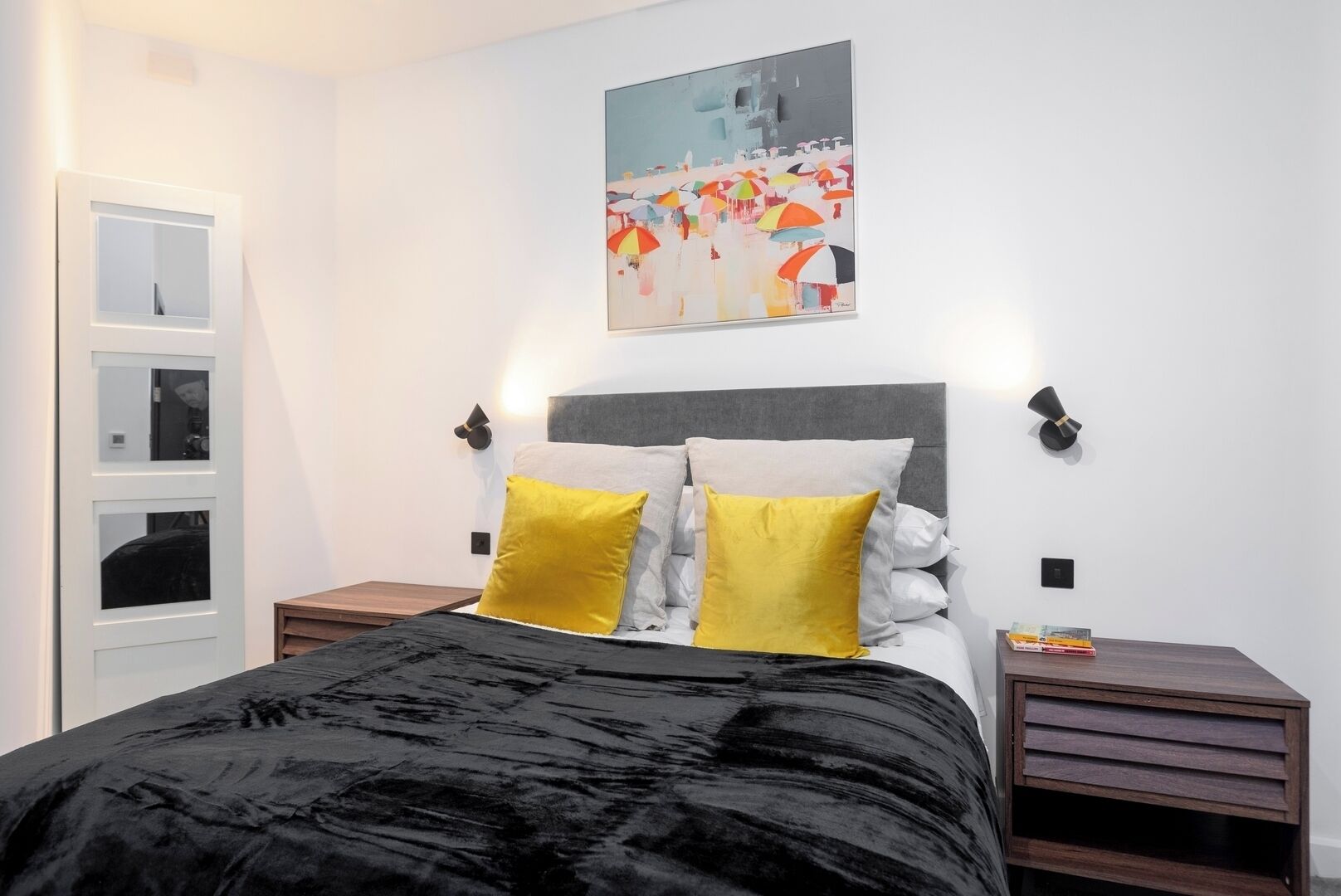 A bedroom with a bed, two bedside tables, a wall-mounted light, and a painting.