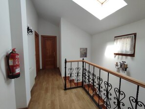 A hallway with a wooden floor, a black metal railing, a fire extinguisher, a wooden door, and a skylight.