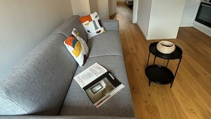 Un salon moderne avec un canapé gris, une petite table d’appoint noire et un magazine posé sur le canapé. Un couloir mène à une cuisine équipée d’un four blanc.