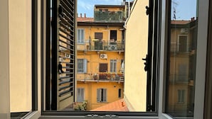 Une fenêtre donnant sur un immeuble jaune, doté de balcons et orné d’une petite plante en pot posée sur le rebord de la fenêtre.