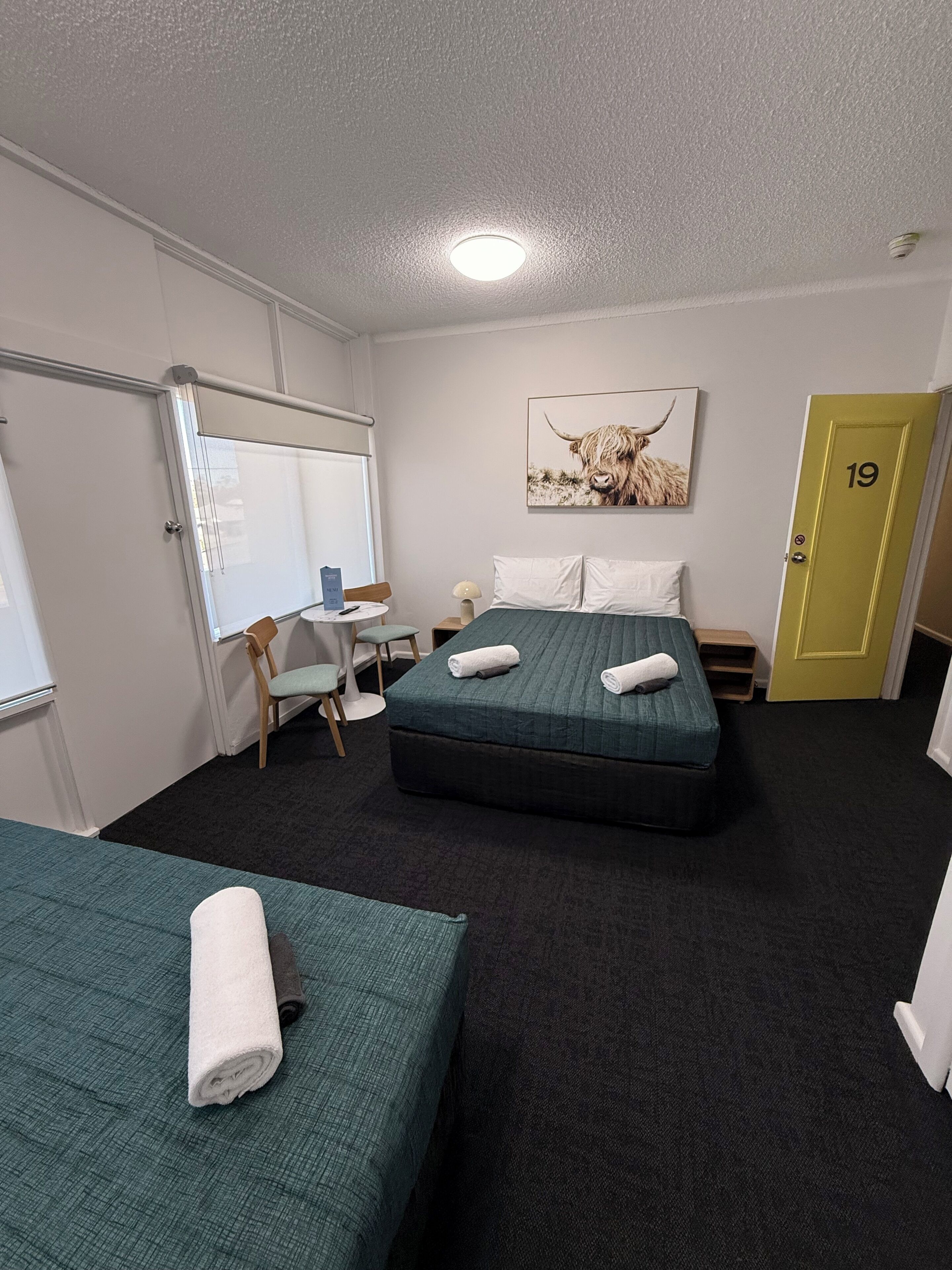 Habitación de hotel con dos camas, un escritorio, una silla, una pizarra y una puerta marcada con el número 19.
