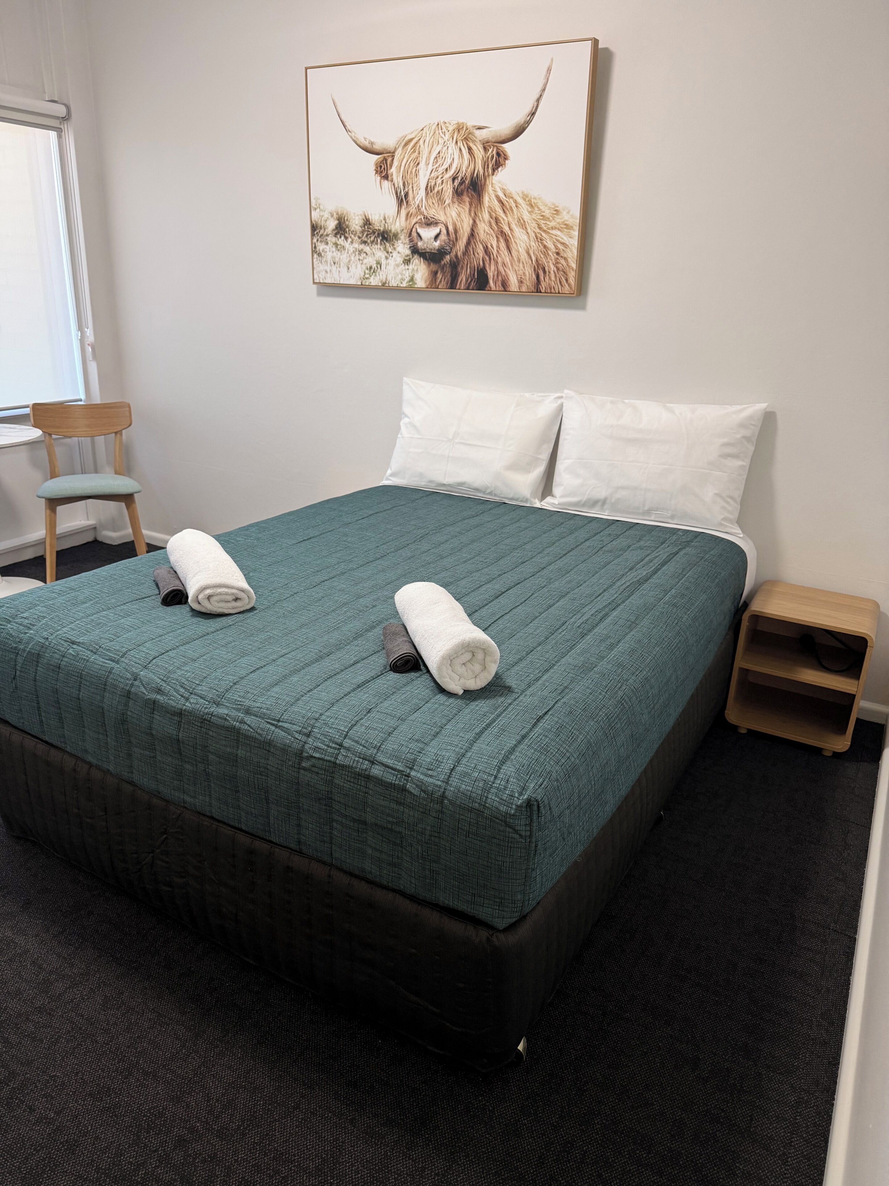 Een hotelkamer met een bed, kussens, een stoel, een nachtkastje en een schilderij van een jak.
