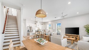 Een moderne keuken met witte kasten, een eethoek met een houten tafel en stoelen, een trap en een woonkamer met een televisie en zitplaatsen.