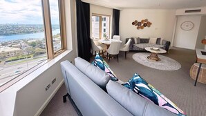 A modern living room with a large window, a sofa, a dining table, and a view of a cityscape and water. - Fancy home with water view in the rocks (The Rocks)
