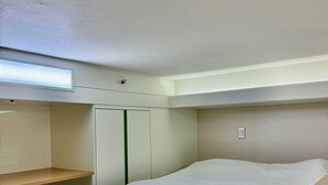 A compact hotel room with a single bed, a small desk, a shelf, and a wardrobe.
