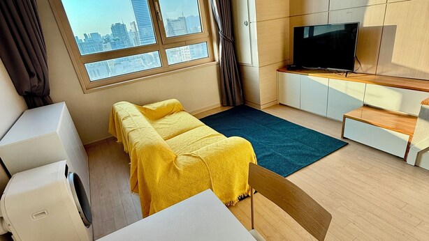 A modern hotel room with a large window, a flat-screen TV, a sofa set, a table, and a washing machine.