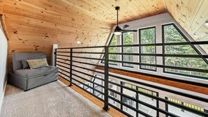 A loft bedroom with a sloped ceiling, a grey sofa, and large windows offering a view of the outdoors. - The Chalet - A Frame in the Overlook, close to Saddleback! (Rangeley)