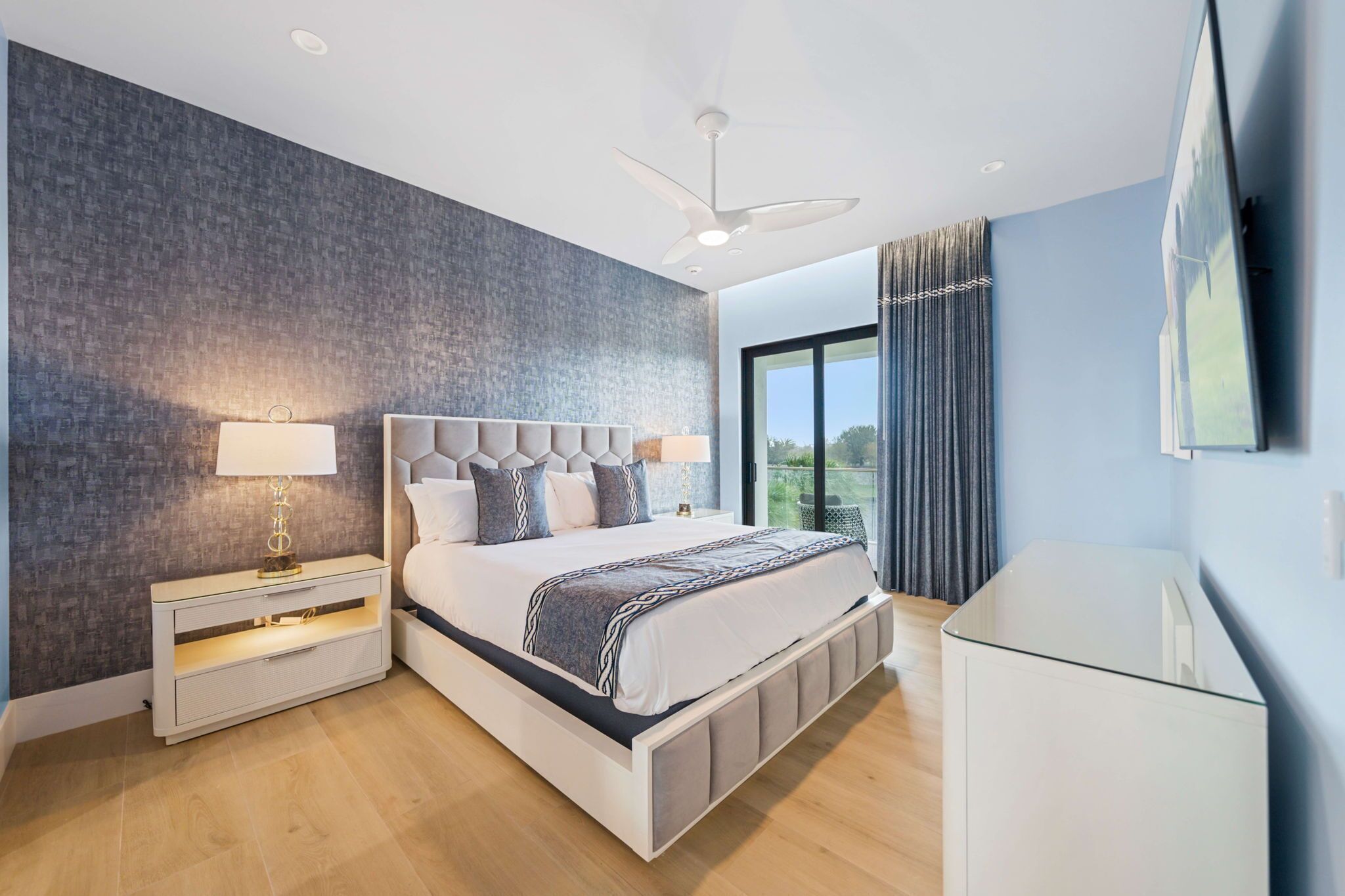 A modern bedroom with a large bed, a nightstand with a lamp, a ceiling fan, and a TV on the wall.