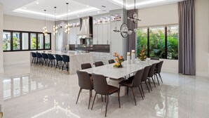 Une cuisine moderne dotée d’un îlot central, d’un coin repas avec une grande table et des chaises, et offrant une vue sur la verdure grâce à de grandes fenêtres.