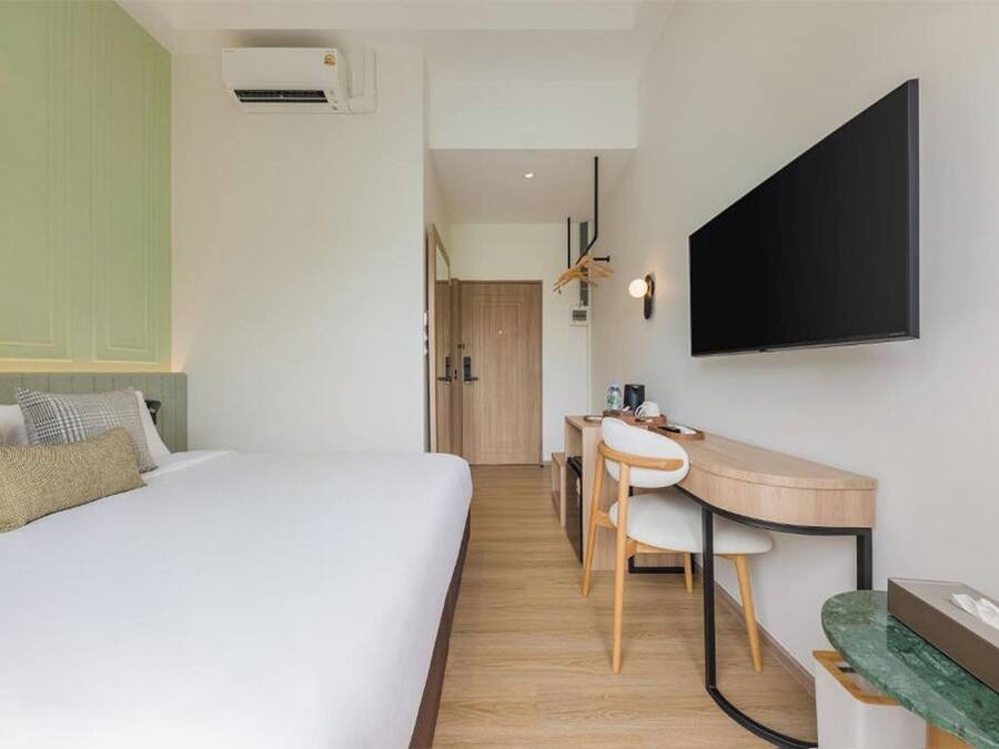 A hotel room with a bed, desk, chairs, a flat-screen TV, and an air conditioning unit.