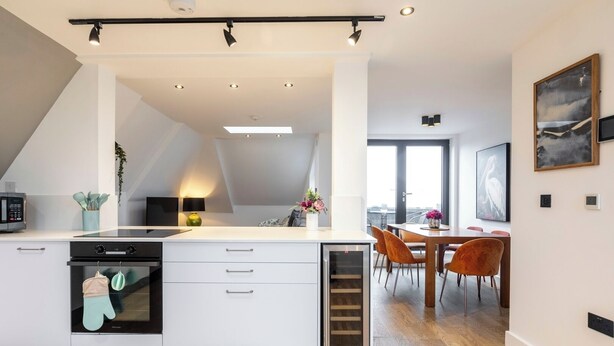 A modern kitchen with a built-in oven, a dining area with a wooden table and orange chairs, and a large window providing natural light.