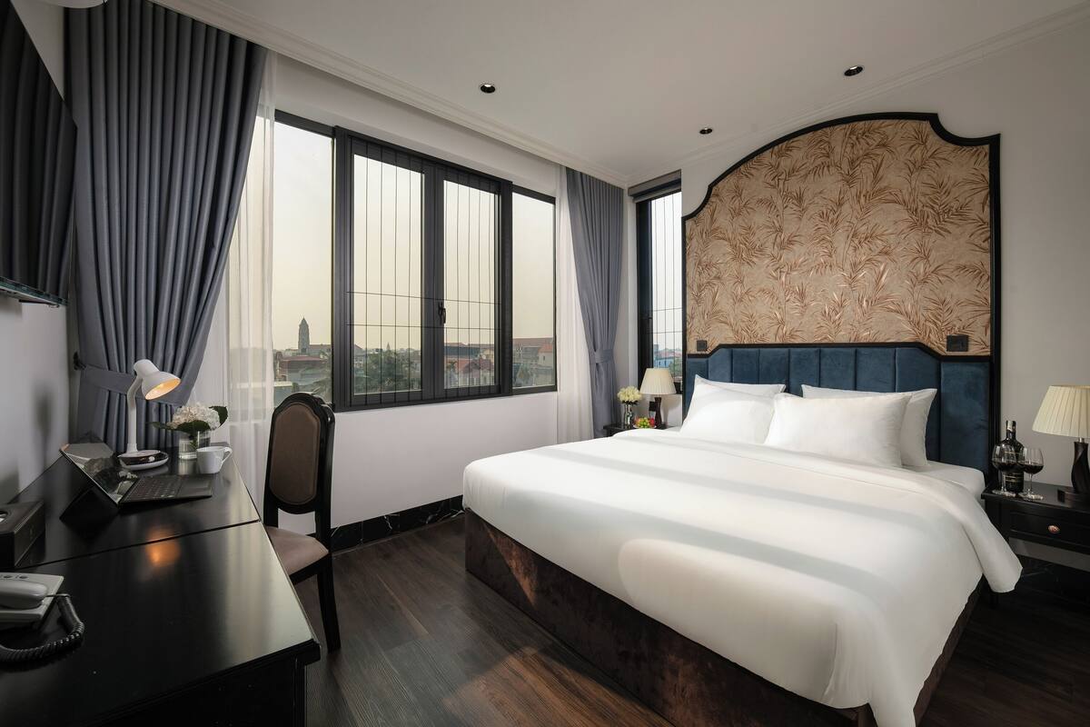 Une chambre d’hôtel avec un grand lit, un bureau avec une lampe, une chaise et une vue sur la ville grâce à de grandes fenêtres.