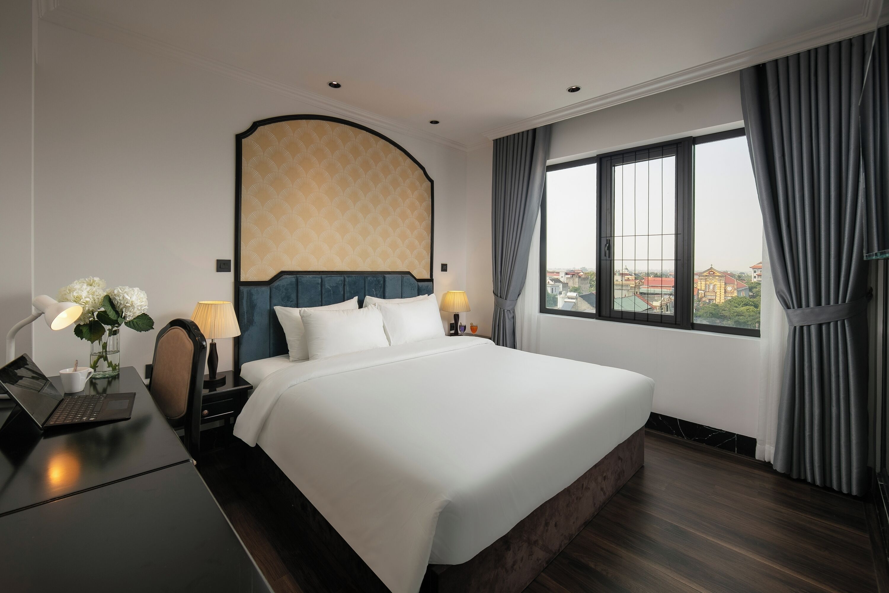 Une chambre d’hôtel avec un grand lit, un bureau avec un ordinateur portable, un vase de fleurs et une vue sur les bâtiments par la fenêtre.