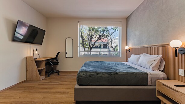 Un dormitorio moderno con cama, escritorio, silla, televisor y ventana con vistas a árboles y edificios.