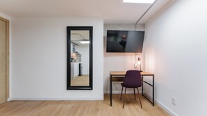 Ein Zimmer mit Holzboden, einem Schreibtisch mit Stuhl, einem an der Wand montierten Fernseher und einem Spiegel.
