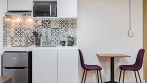 Eine kompakte Küche mit Mikrowelle, Kaffeemaschine und kleinem Kühlschrank. Zwei violette Stühle und ein Holztisch sind ebenfalls vorhanden.