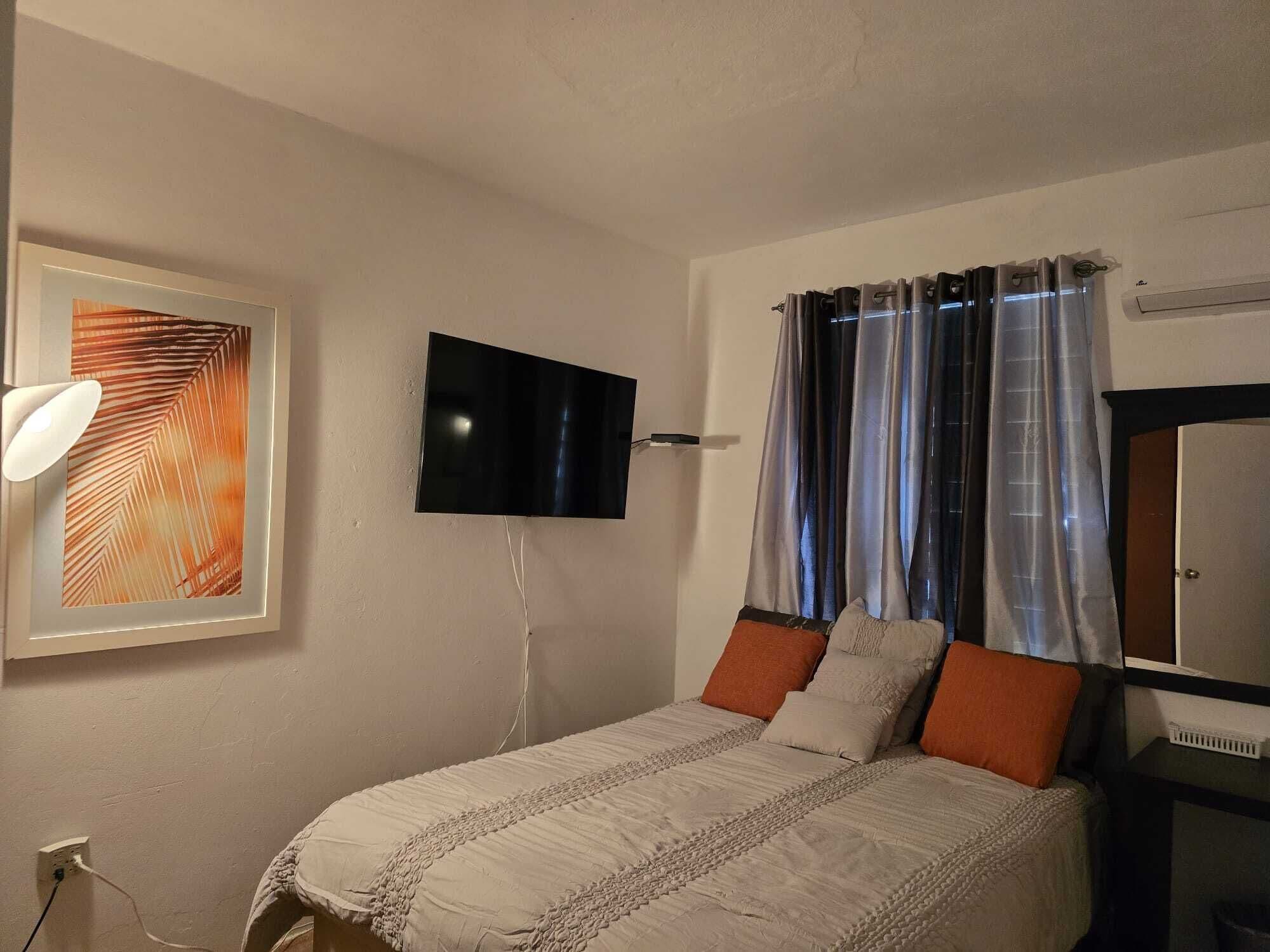 Een slaapkamer met een bed, een flatscreen televisie aan de muur, een ingelijst kunstwerk en een raam met gordijnen.