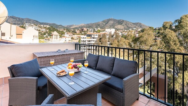 A balcony with wicker furniture, a table with fruit and drinks, and a view of a town and mountains. - Mountain Views, Pool & Rooftop: Cozy 2BR for 6 REF034 (Fuengirola)
