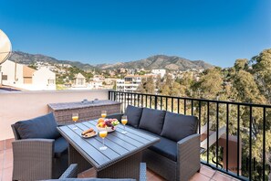 Ein Balkon mit Korbmöbeln, einem Tisch mit Obst und Getränken sowie Blick auf eine Stadt und Berge.
