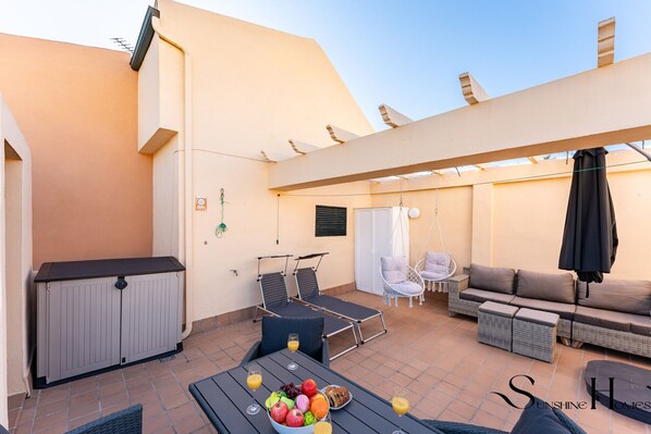 Eine sonnendurchflutete Terrasse im Freien mit Esstisch für zwei Personen, einer Couch und einem Schaukelstuhl.