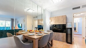 A modern dining area with a wooden table set for four, surrounded by chairs. A fruit bowl and glasses of orange juice are on the table. - Mountain Views, Pool & Rooftop: Cozy 2BR for 6 REF034 (Fuengirola)