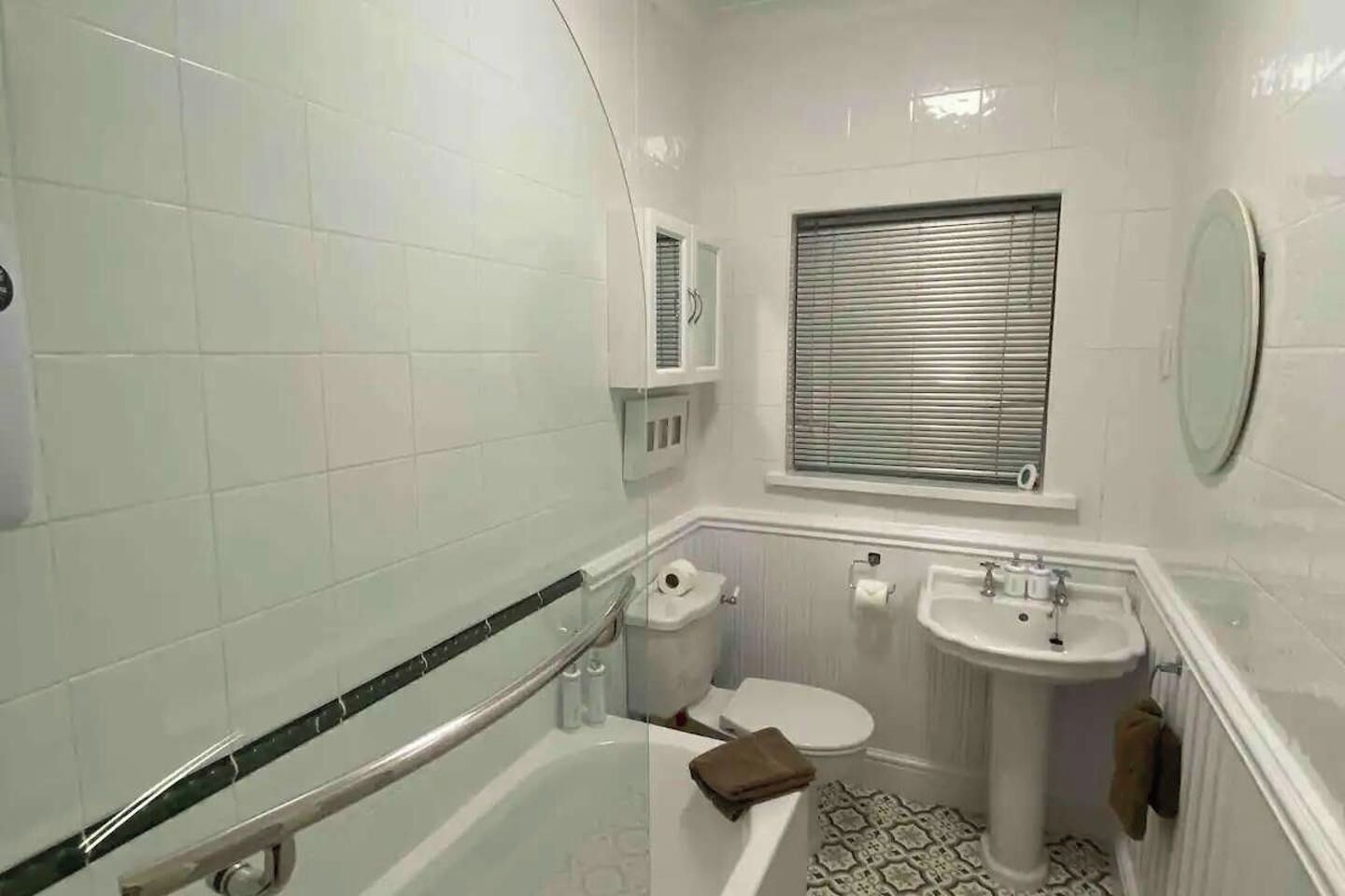 A bathroom with white tiles, a bathtub, a toilet, a sink, and a window with blinds.