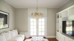 A spacious living room with a white sofa, a large ottoman, a chandelier, a television on a white cabinet, and a large window with a view of greenery.
