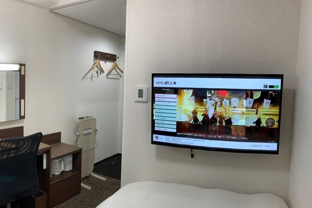 Ein Hotelzimmer mit einem Bett, einem Schreibtisch mit Stuhl, einem an der Wand montierten Fernseher und einem Spiegel über dem Schreibtisch.