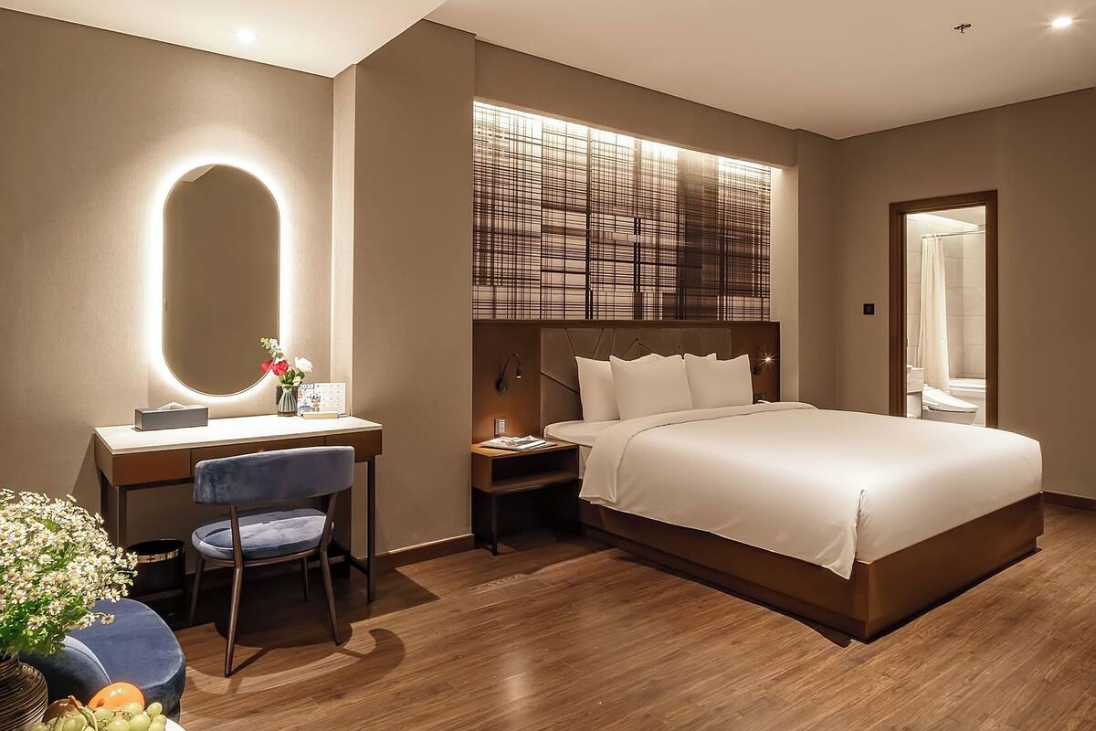 A modern hotel room with a large bed, a desk with a chair, a vanity with a mirror, and a small table with a vase of flowers.