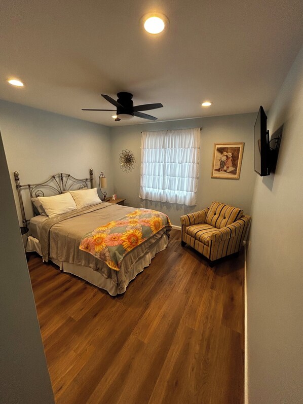 Un dormitorio con cama, una silla, ventilador de techo, ventana con cortinas y televisor montado en la pared.
