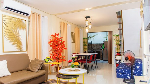 A modern living room with a sofa, a small round table, and a dining area with chairs. There is a fan and a decorative plant in the room.