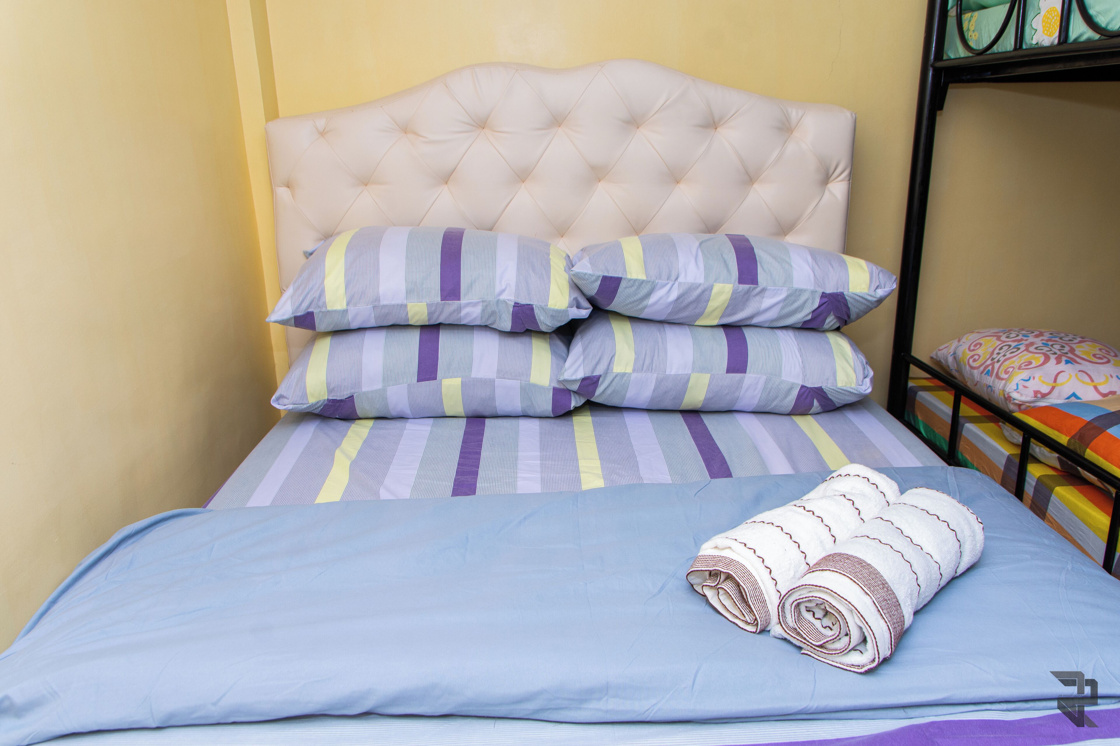 Una cama con cabecera capitonada, almohadas a rayas y una toalla enrollada.