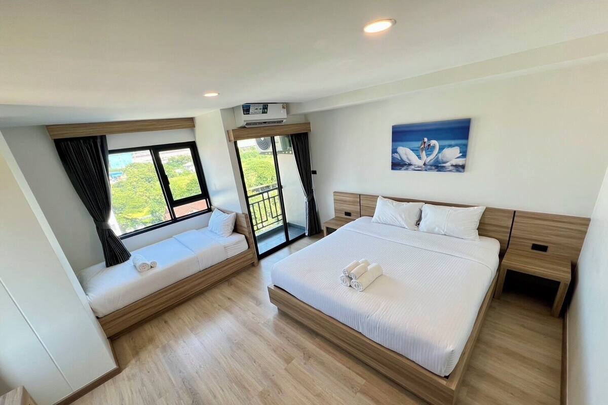 A modern hotel room with a large bed, a smaller sofa, a balcony with a view, and an air conditioning unit.