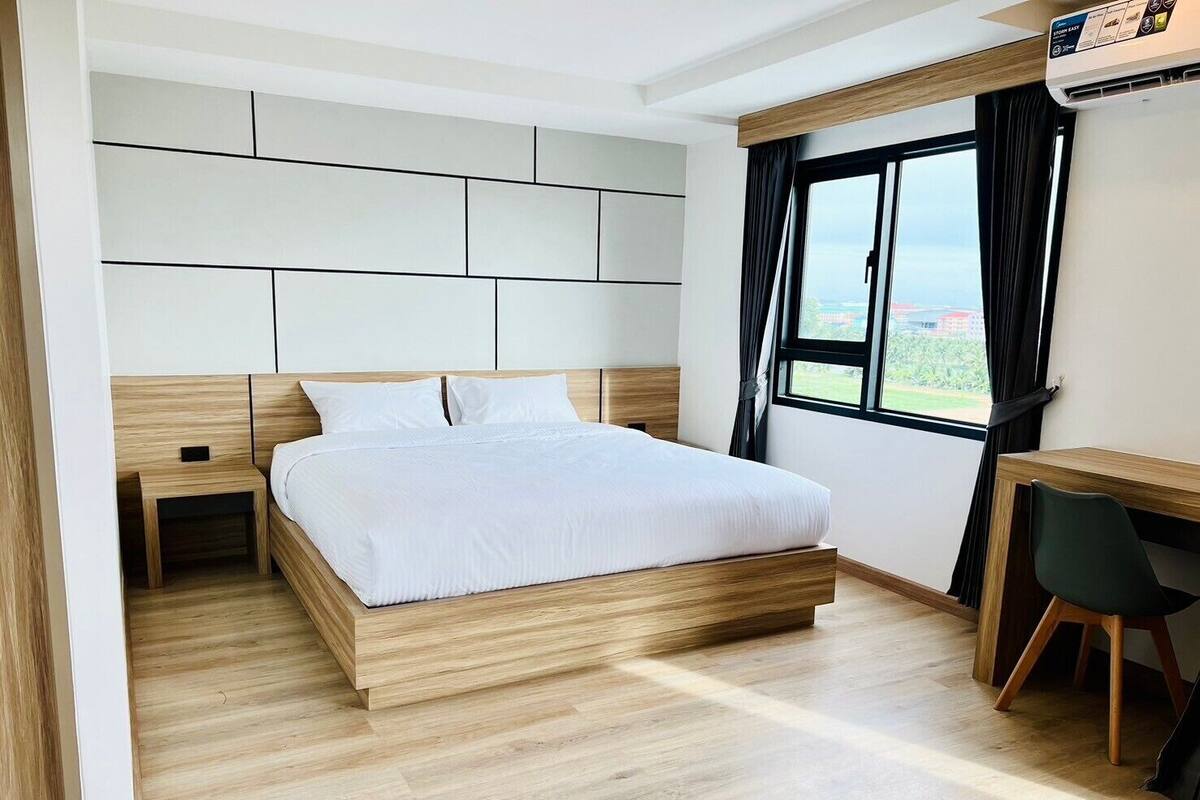 A modern bedroom with a wooden bed, a small bedside table, a desk with a chair, and a window with curtains.