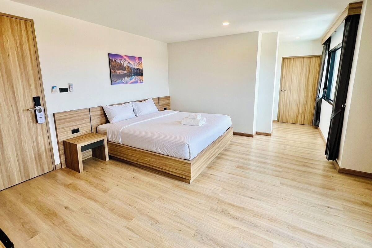 A modern hotel room with a wooden floor, a large bed with white linens, a nightstand, a wall-mounted painting, and a door with a keycard lock.